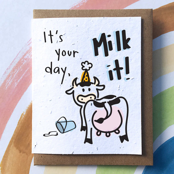 A white card on a brown envelope. On the card there is a cow wearing a yellow birthday hat standing next to a spilt bucket of milk. Above, the text reads "It's your day, Milk it!"