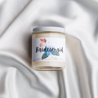 A candle with the word “Bridesmaid” on it on top of a floral design