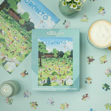 Villager Puzzles | The Six in the Sun | 500-Piece Puzzle for Adults - Turquoise box of Villager Puzzles 500-piece Toronto-themed jigsaw with scattered colorful puzzle pieces and latte on pastel.