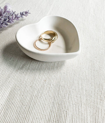 Mini Heart Dish (no tariffs) - Small white ceramic heart-shaped dish holding gold rings on textured fabric background.