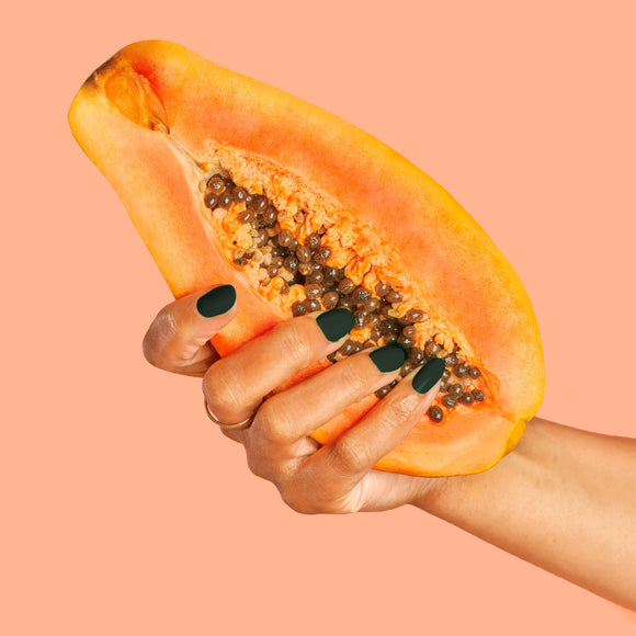 Vegan & 21-free Nail Polish - En Beau Fusil - Hand with dark green vegan nail polish holding halved ripe papaya against peach background.