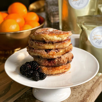 Crispy Cinnamon Doughnut Pancake Mix | Maison Zoe Ford - Stack of golden-brown crispy cinnamon doughnut pancakes dusted with sugar on a white pedestal plate with fresh blackberries.