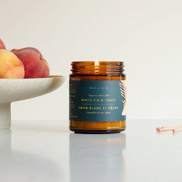 White Fir & Peach Candle - Amber glass candle jar with dark blue label and gold text, sitting on white surface next to white bowl with peaches.