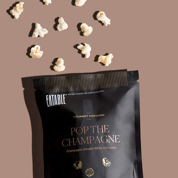 Pop the Champagne - Gourmet Popcorn (Eatable) - Black matte bag of handmade Canadian gourmet popcorn labeled Pop the Champagne with scattered popcorn kernels as unique gift.