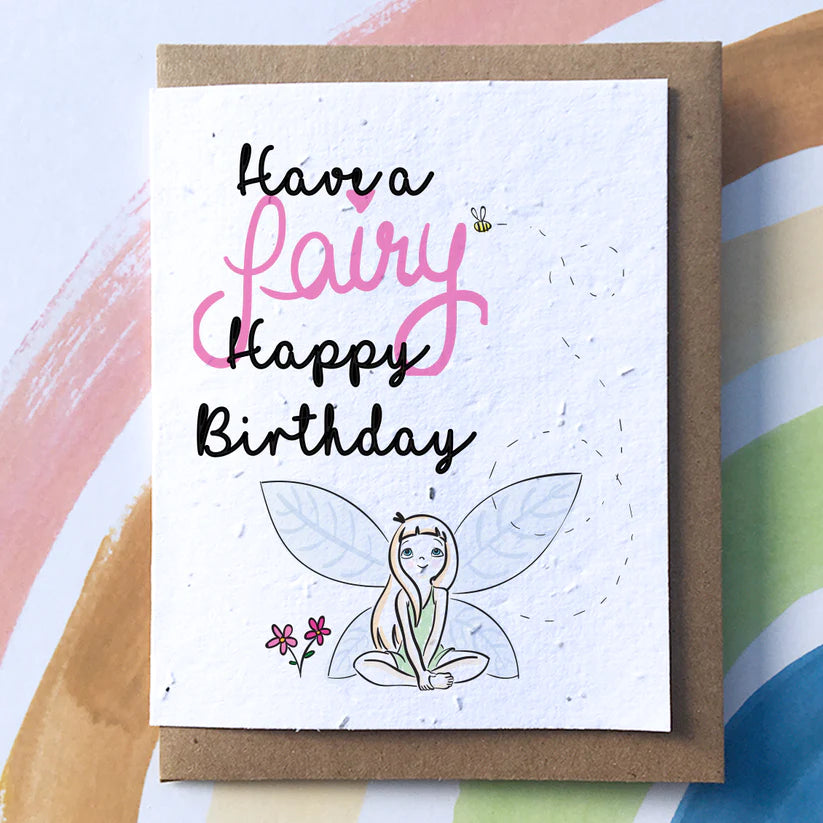 A white card with a brown envelope. On the card reads "Have a fairy Happy Birthday" with the word fairy in pink and the rest of the text in black. Underneath there is a fairy watching a bee fly next to the word fairy.