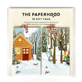 Two gift tag sets next to each other. The tag on the left depicts a brown cottage in a wintry forest. There are a pair of red skies and a tree stump with an axe next to the cottage. The tag on the right depicts two people cross-country skiing through a wintry forest. 