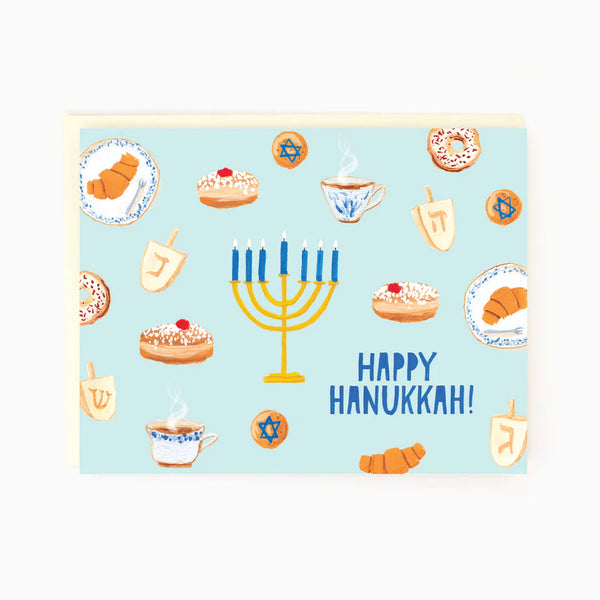 A light blue card with sufganiyat, challah, rugelach, babka and dreidels surrounding a lit menorah. In the bottom right corner reads in blue 