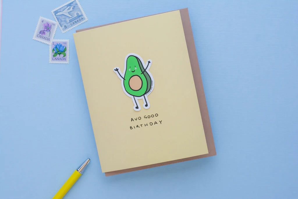 A light brown card with a brown envelope and a green illustrated avocado in the middle with the text "Avo good birthday" underneath