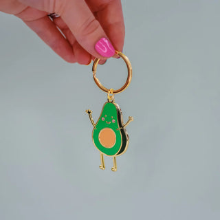 A person with pink nails holds a gold keychain with a green illustrated smiling avocado on it