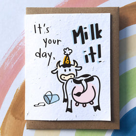 A white card on a brown envelope. On the card there is a cow wearing a yellow birthday hat standing next to a spilt bucket of milk. Above, the text reads "It's your day, Milk it!"