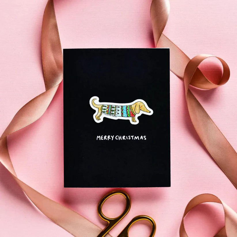 A black card with a dachshund dog in the center, wearing a Christmas sweater. Underneath reads "MERRY CHRISTMAS" in white