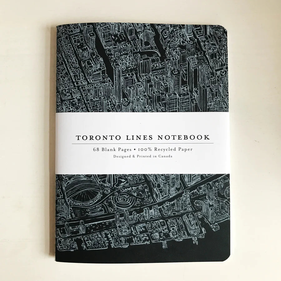 A black notebook with a detailed, fine-line white illustration of Toronto from a birds-eye view. A removable label sleeve reads "Toronto Lines Notebook, 68 blank pages, 100% recycled paper, Designed & Printed in Canada"