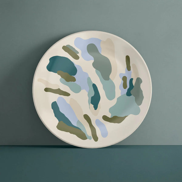 A cream coloured plate with abstract splashes of olive green, light blue, turquoise and cream against a dark blue-green background