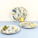 2 plates standing up against a pastel yellow wall with a pear in front of them. 2 more plates are holding popcorn and a kiwi cut in half
