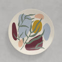 A cream coloured plate with abstract splashes of maroon, salmon, mustard yellow and light blue with green leaves painted on top against a grey background