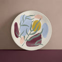 A cream coloured plate with abstract splashes of maroon, salmon, mustard yellow and light blue with green leaves painted on top against a faded maroon background