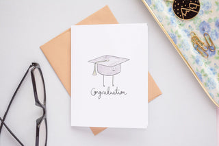 A white card with a blue graduation cap with a smiley face in the middle. Underneath in black cursive reads 