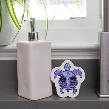 Eco Sponges - Purple and black turtle-shaped sponge with intricate Indigenous-style design next to beige soap dispenser.