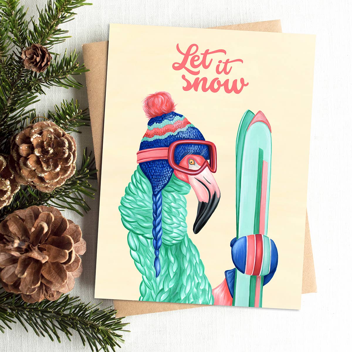 A white card with a flamingo wearing a winter hat and scarf, and wearing ski goggles and holding skiis and the words "Let it Snow"