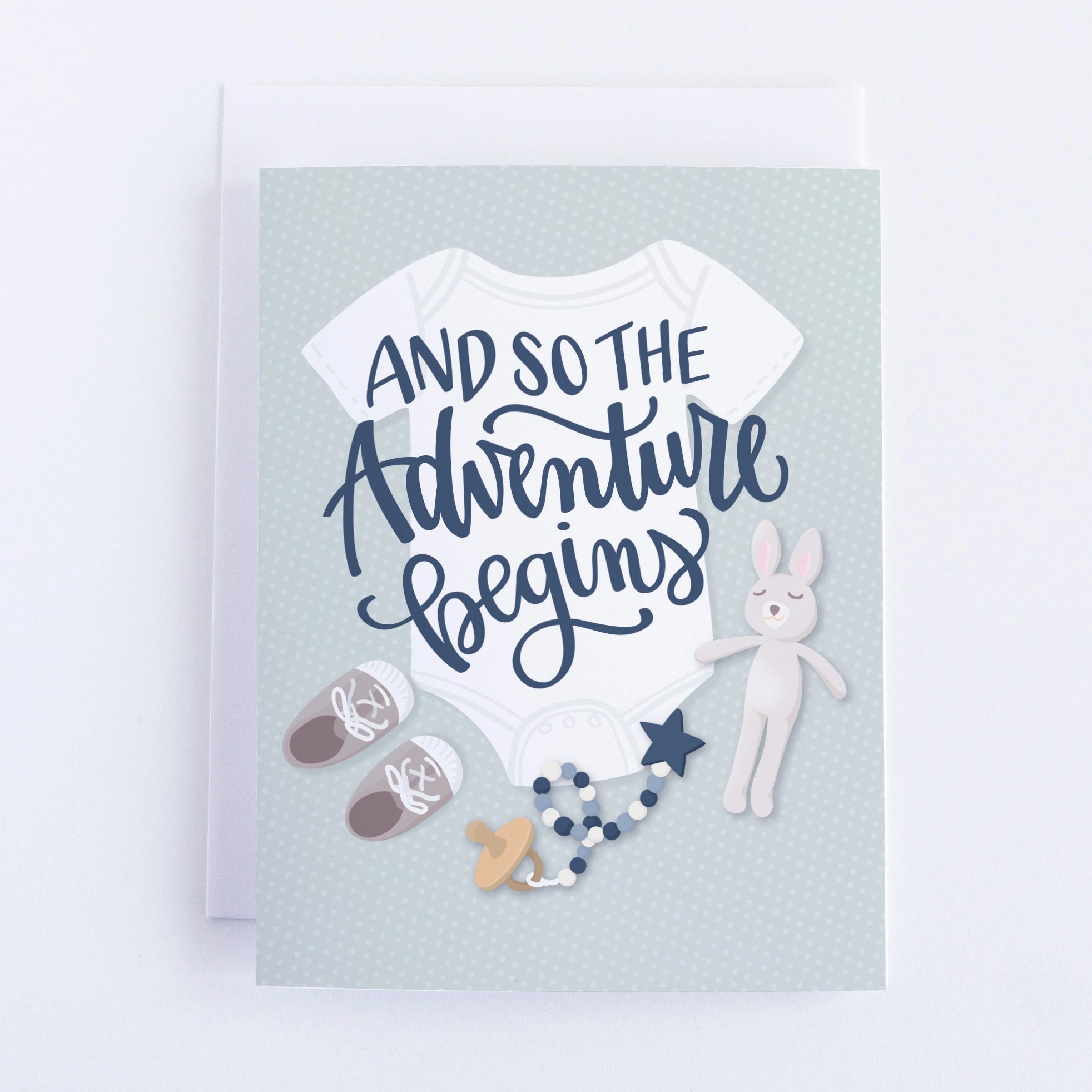 Grey blue background with a white baby jumper, a bunny, grey baby sneakers, a blue pacifier and a plush bunny with the message "And so the adventure begins" in navy