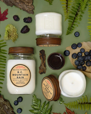A glass jar with a brown, rustic lid with the candle inside laid out next to leaves, berries, 2 other candles and bark on a green background