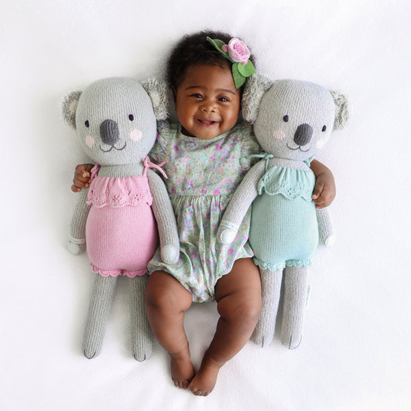 2 koalas on either side of a baby sleeping, the one on the left wearing a pink dress and the one on the right wearing a teal dress