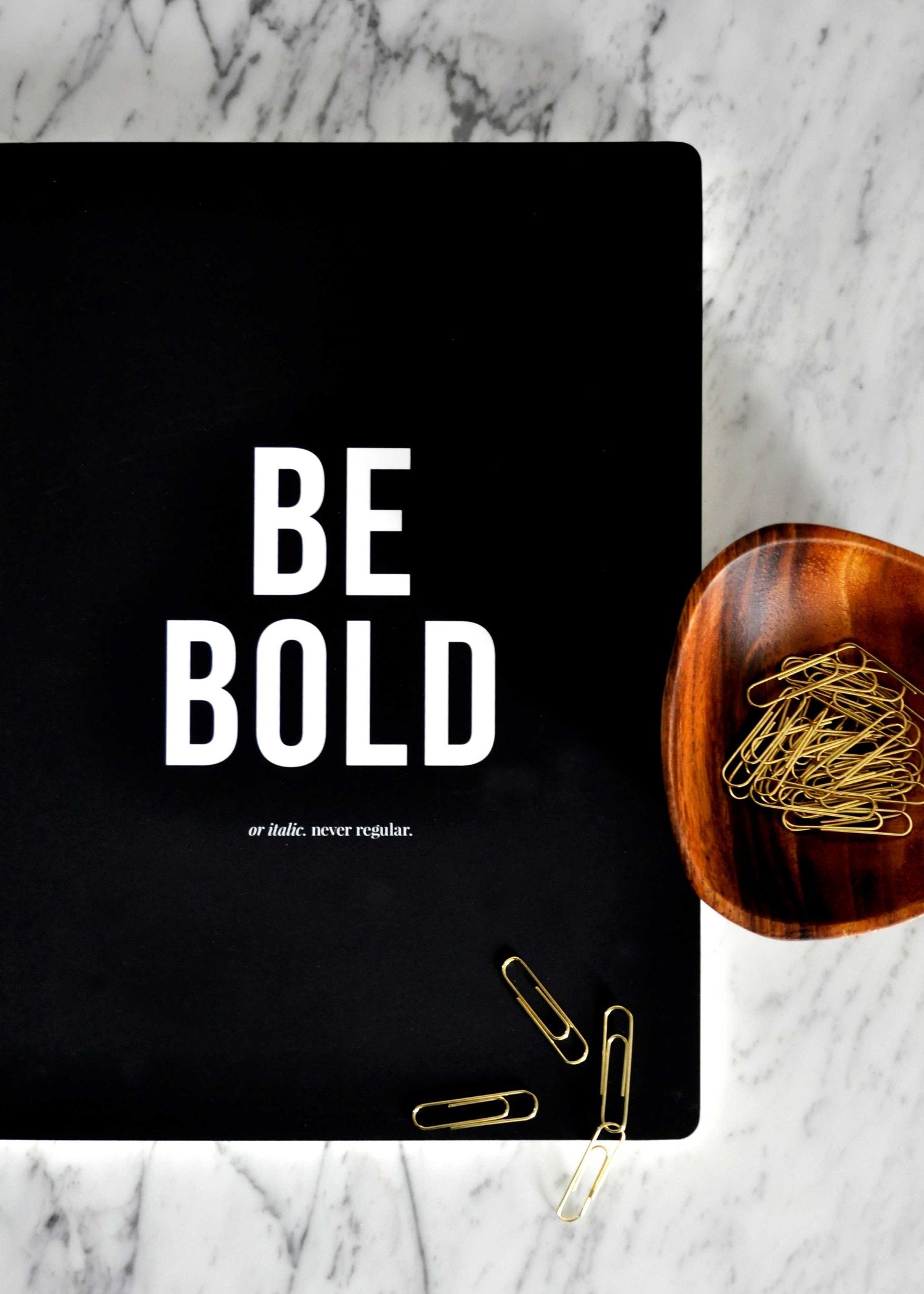 A matte black notebook with white text reading "Be Bold or italic. never regular" next to a wooden bowl of gold paperclips